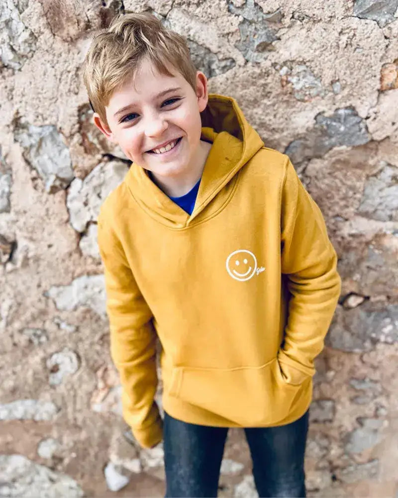 Lachender Junge im gelben Hoodie mit Smiley-Aufdruck, vor Steinmauer stehend