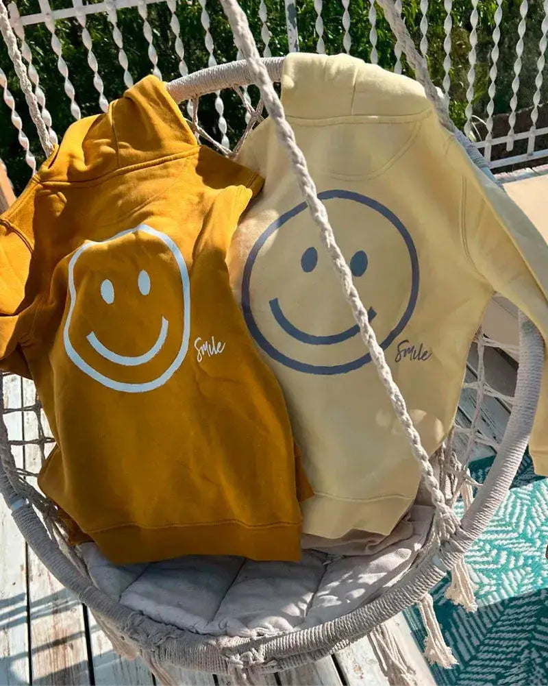 Zwei Kinderhoodies in Gelb und Ocker mit großem Smiley-Aufdruck und „Smile“-Schriftzug auf Hängesessel