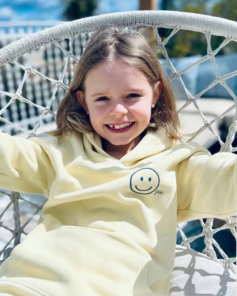 Mädchen im pastellgelben Hoodie mit Smiley-Aufdruck, entspannt im Outdoor-Hängesessel