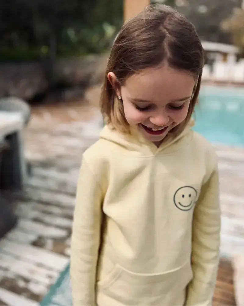 Mädchen mit pastellgelbem Kinder-Hoodie mit Smiley-Aufdruck steht draußen am Pool