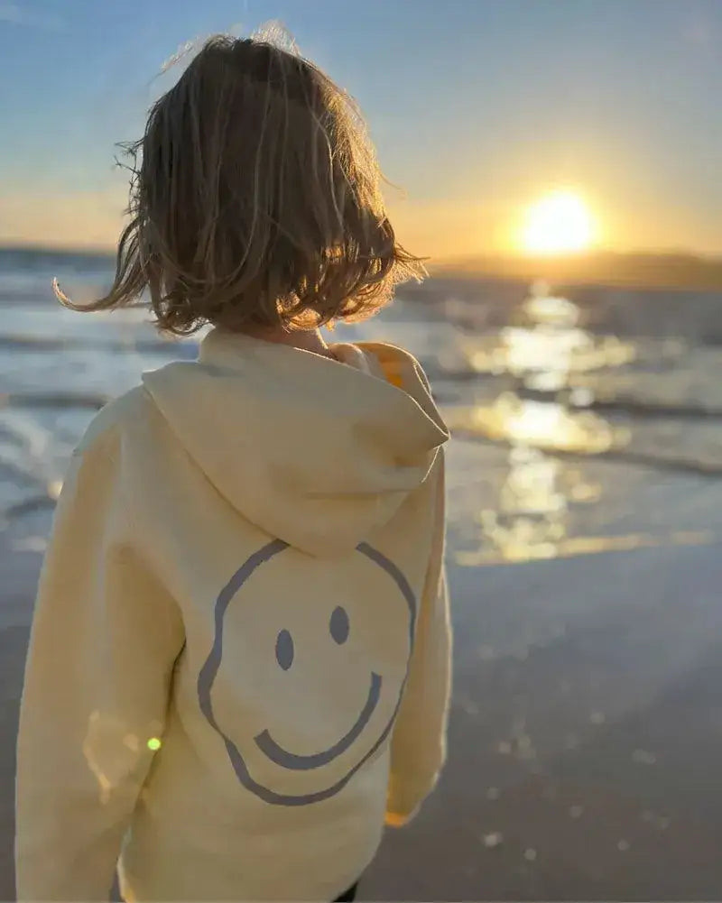 Kind am Strand im Sonnenuntergang mit pastellgelbem Kinder Hoodie und großem Smiley Aufdruck