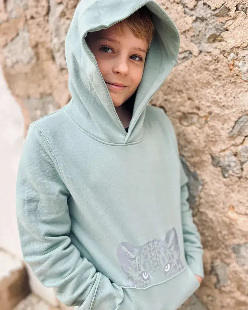 Junge im mintgrünen Bio-Kinderhoodie mit Leoparden-Print an der Wand stehend.