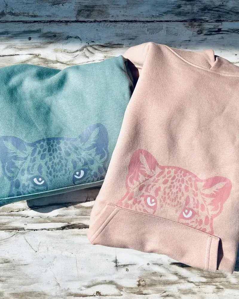 Zwei nachhaltige Kinder Hoodies in Mint und Pfirsich mit Leoparden-Motiv auf Holzuntergrund