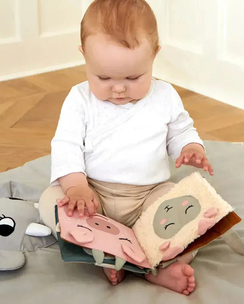 Baby sitzt auf Boden und blättert in nachhaltigem Stoffbuch mit Tiermotiven Schwein und Schaf.