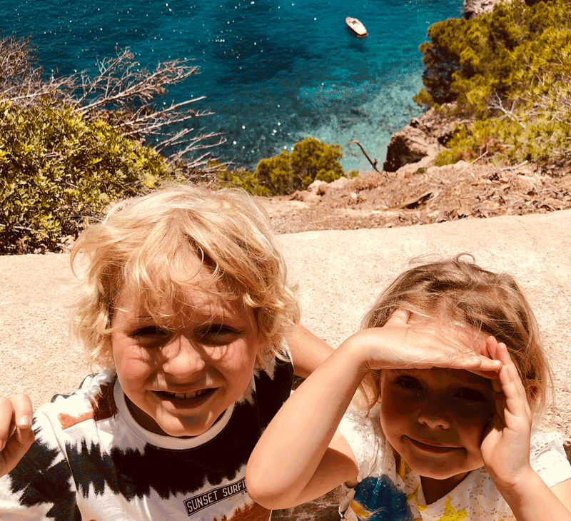 Mädchen und Junge schauen uns an. Sie stehen auf einem Berg. Im Hintergrund sieht man das schöne, hellblauschimmernde Meer.