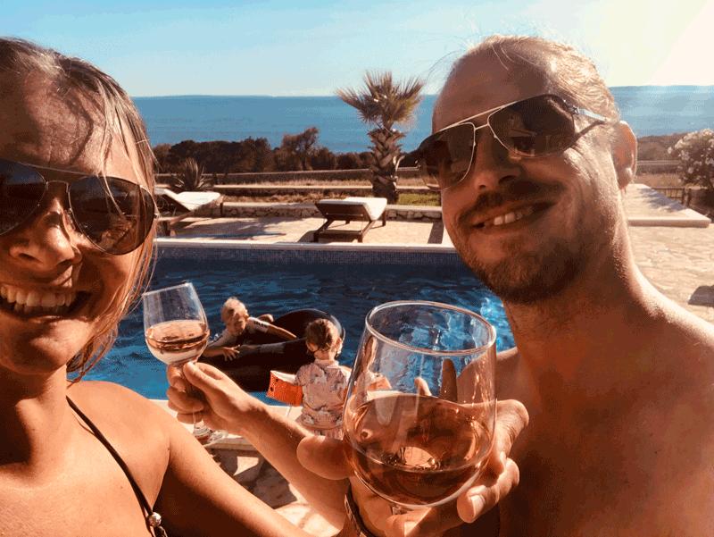 Familie trinkt auf einen wundervollen Tag am Pool mit Ausblick auf das Meer. Eine Palme steht im Hintergrund und die Kinder spielen mit einem Schwimmring im Wasser.