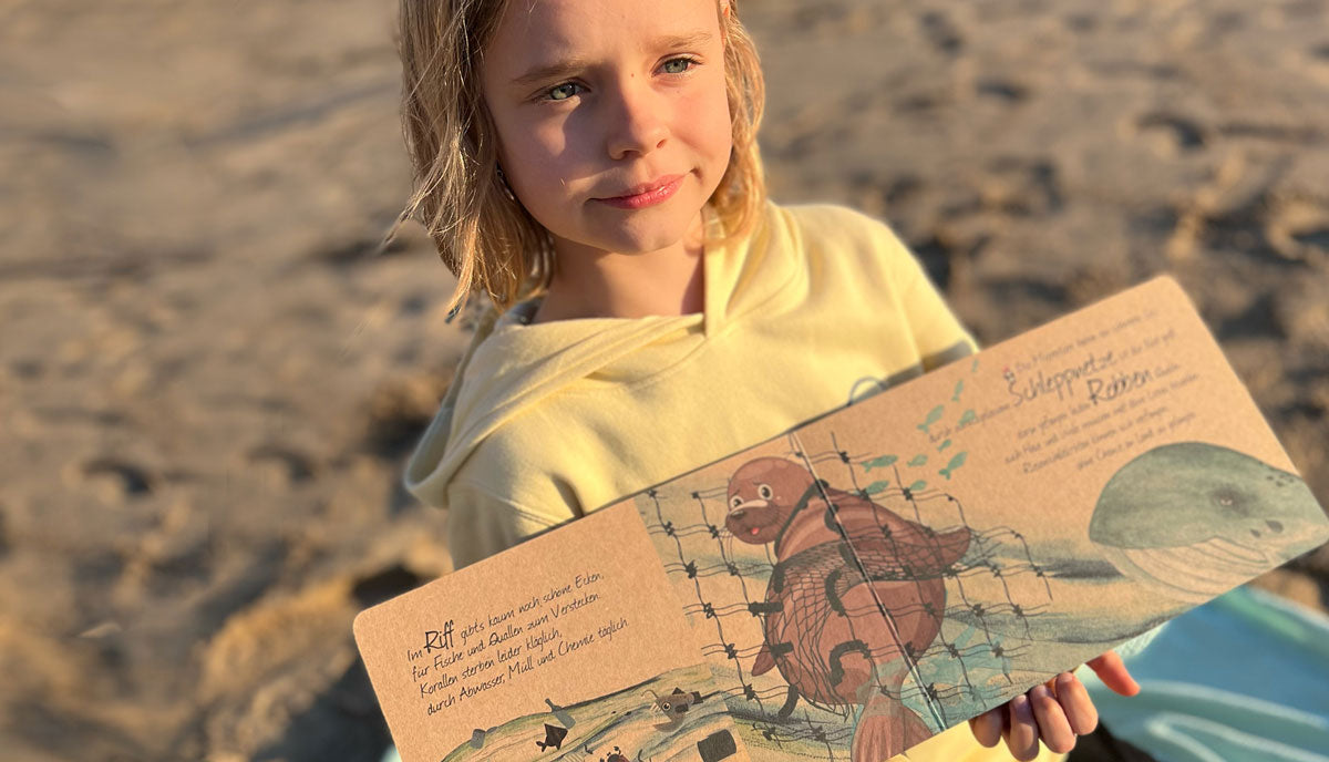 Mädchen zeigt eine Seite des Kinderbuchs "Der Wal auf seiner Reise". Auf der Seite sieht man eine Robbe, die in einem Netz gefangen ist. Ein Wal weint daneben.