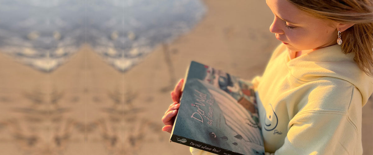 Ein Mädchen steht am Strand und schaur sich ein Buch über den Müll im Meer an. Auf dem Titel ist ein Wal, der weint.