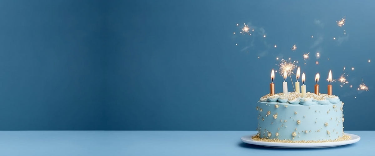 Man sieht eine blaue Geburtstagstorte mit Kerzen, die angezündet sind. Eine Wunderkerze sprüht Funken in die Luft. Der Hintergrund ist blau und die Oberfläche, auf dem der Kuchen steht hellblau. 